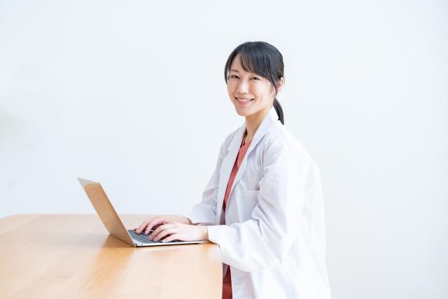 ノートパソコンの前でほほえむ白衣の女性医師